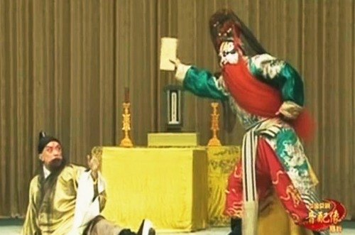 京剧《打金砖》剧照:太庙一场,供桌设神主牌位与烛台,一角色跌坐惊退,净角(红黑脸谱、红满髯、绿蟒)立而举牌(央视网《京剧音配像〈打金砖〉》文内所载)