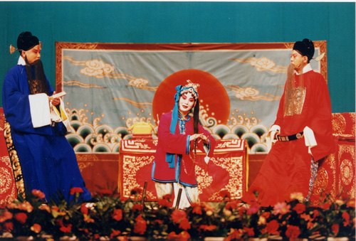 京剧《玉堂春》剧照:三堂会审一场,苏三(罪衣、鱼枷)与众官员同台(国家京剧院官网剧目页所载)