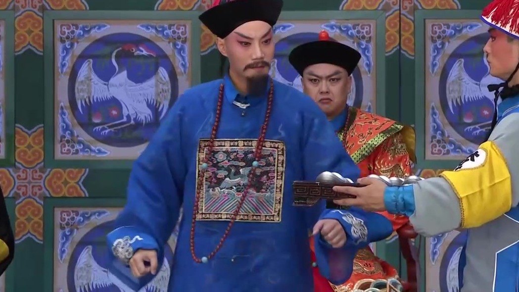 京剧《瘦马御史》演出照:差役托盘呈银锭、御史(蓝官衣、朝珠、黑三髯)伸手探看,红蟒官员在后——查库一节官场场面