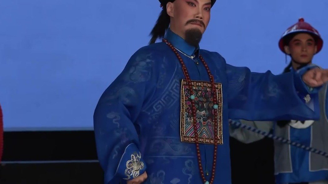 京剧《瘦马御史》演出照:老生扮御史(宝蓝官衣、补子、朝珠、黑三髯)作指认身段,身后差役牵铁链