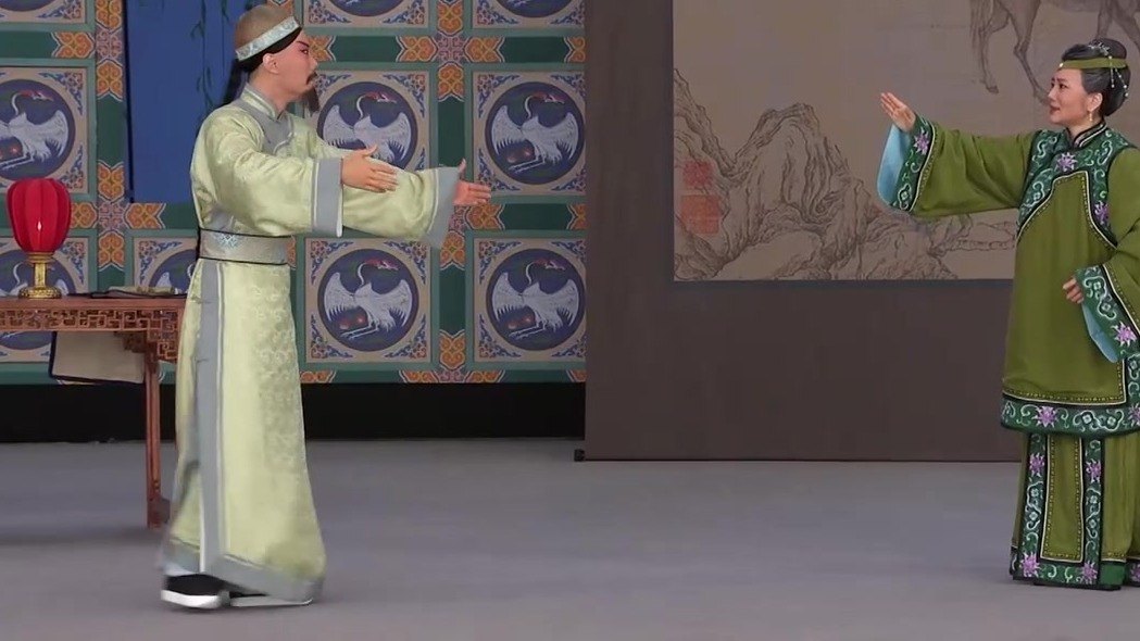 京剧《瘦马御史》演出照:老生(浅豆绿褶袍、髯口)与旦角对戏,舞台设桌灯、鹤纹屏与山水画屏