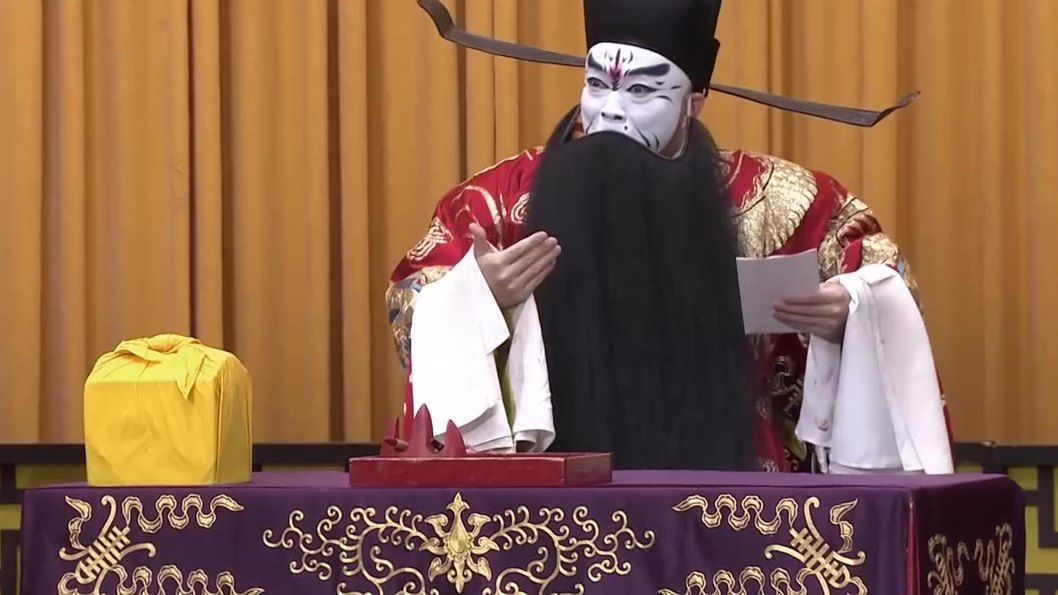 京剧《群英会·借东风·华容道》第四场演出照:红蟒、白脸黑满髯的净角立于桌案后(央视《CCTV空中剧院》)