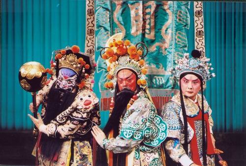 京剧《群英会》剧照:三人同场亮相——左为勾脸净角(黑髯、绒球珠饰硬盔、黑底金绣戏衣,持金色瓜形砌末),中为红脸长髯角色(绿底金绣戏衣),右为俊扮无髯年轻男角色(浅色绣花戏衣、系红带);背景为湖蓝幕布与绘龙纹竖幅守旧(国家京剧院官网剧目页所载,未标注场次)