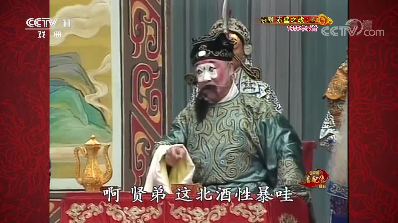 CCTV-11《中国京剧音配像精粹》京剧《赤壁之战》(上,1959年录音)播出画面:丑角扮相人物坐于桌案后举壶劝酒,旁立扎靠武将,底部字幕“啊贤弟 这杯酒性暴哇”